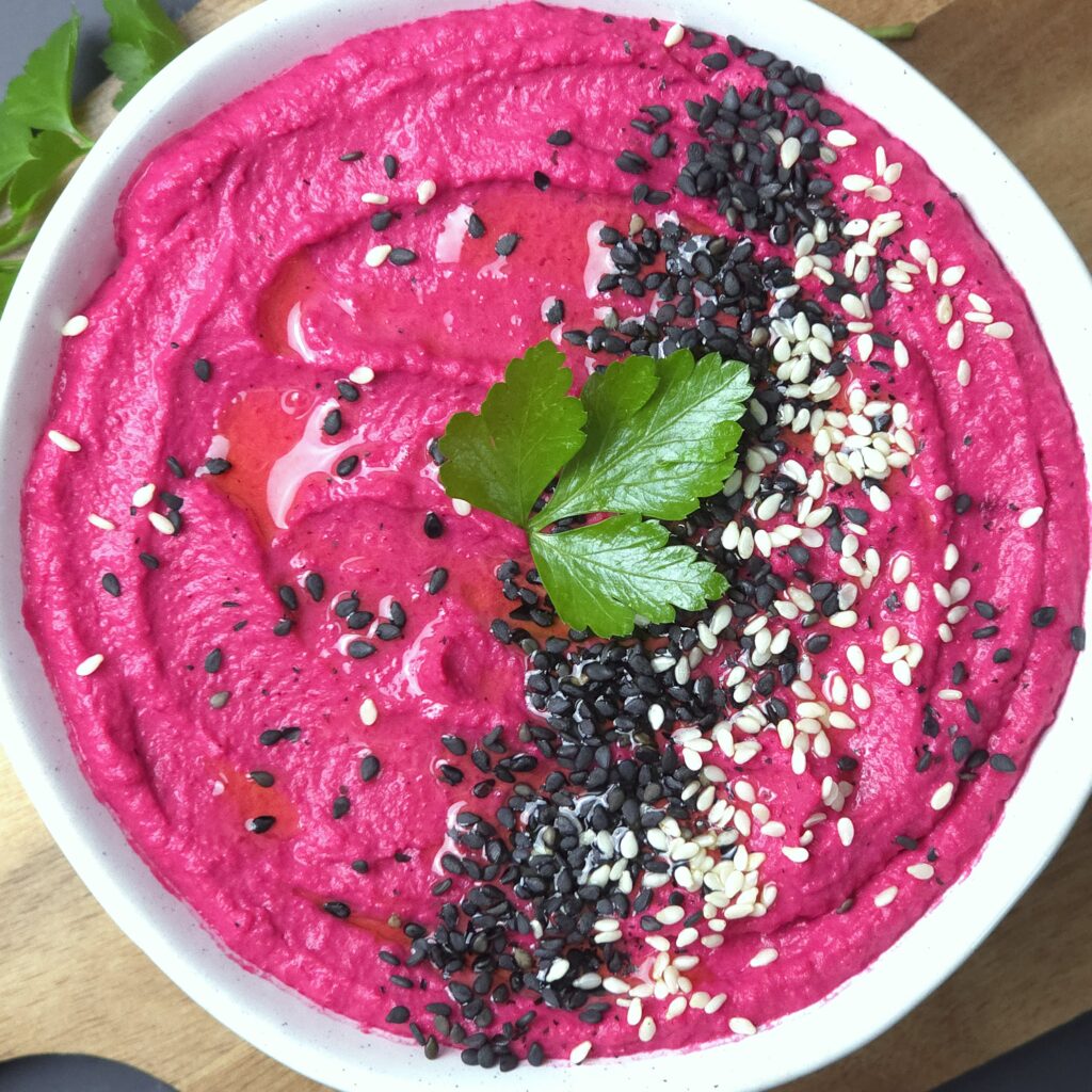 beetroot hummus recipe in a white stoneware bowl, sprinkled with black and white sesame seeds