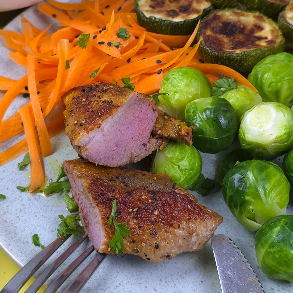 cutted in a half roasted duck breast in a plate next to steamed brussels sprouts, roasted courgette, carrot salad