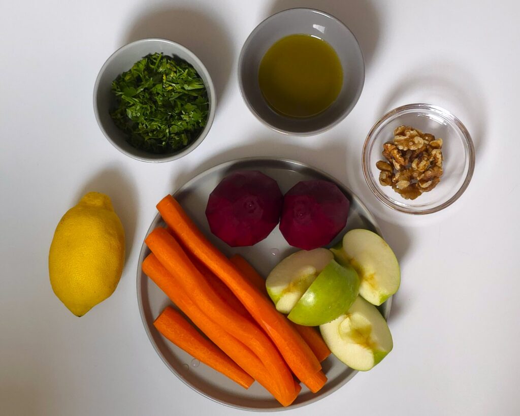 beetroots, carrots, apple in a plate next to lemon, freshly chopped parsley, olive oil and walnuts in bowls