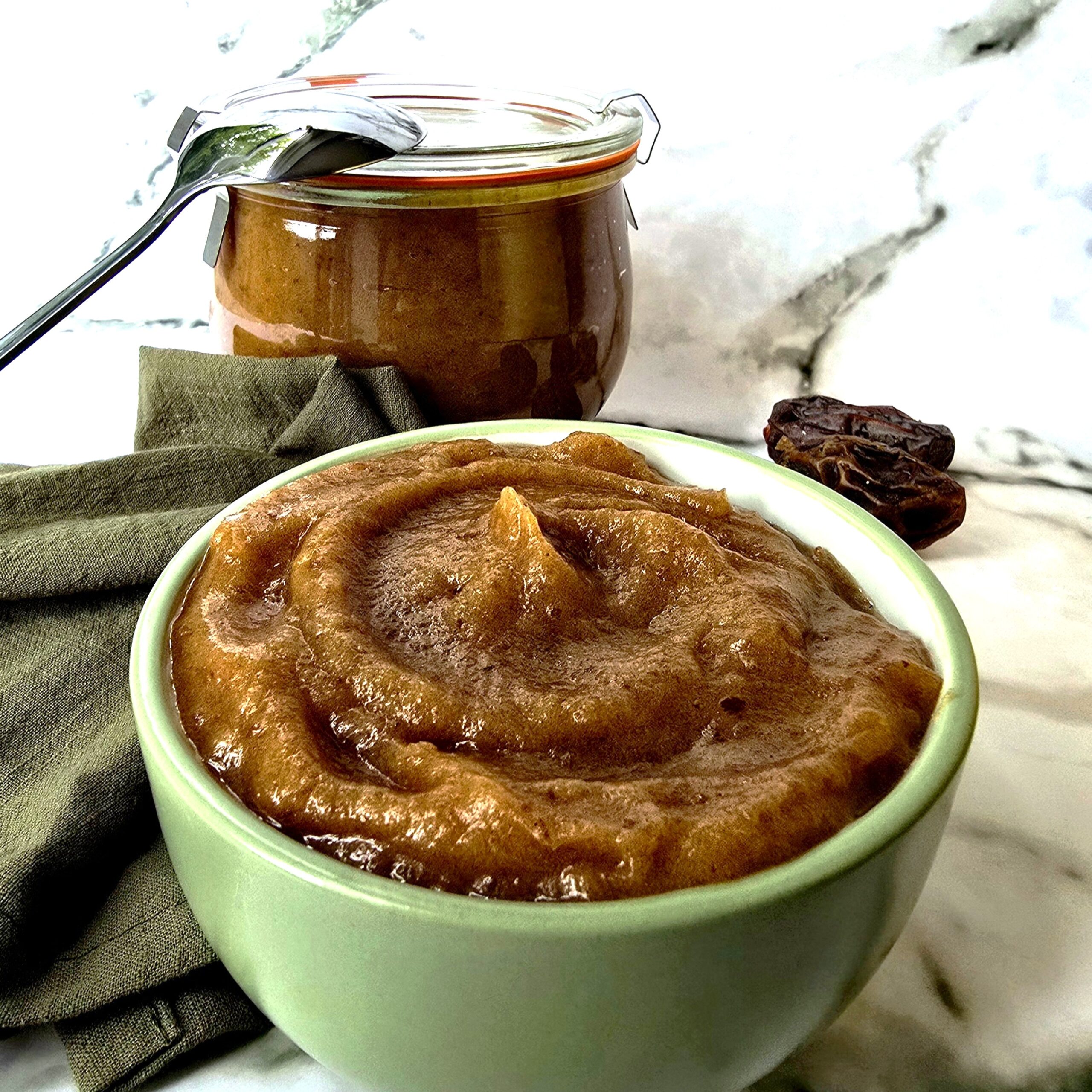a green bowl with homemade date paste and jar with date paste at the background and spoon