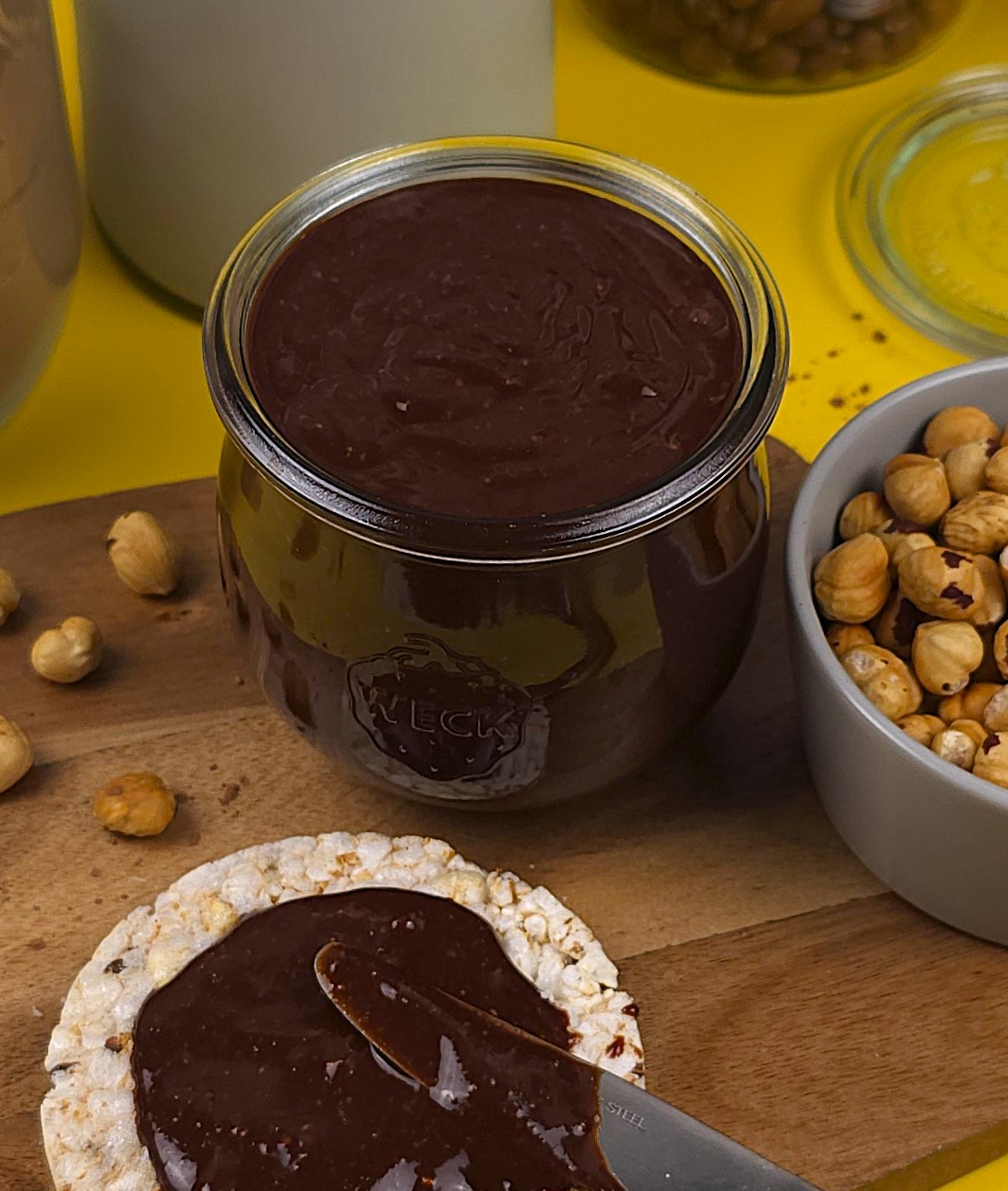 a jar with a homemade nutella next to a rice cake topped with hazelnut spread, and a bowl with roasted hazelnuts