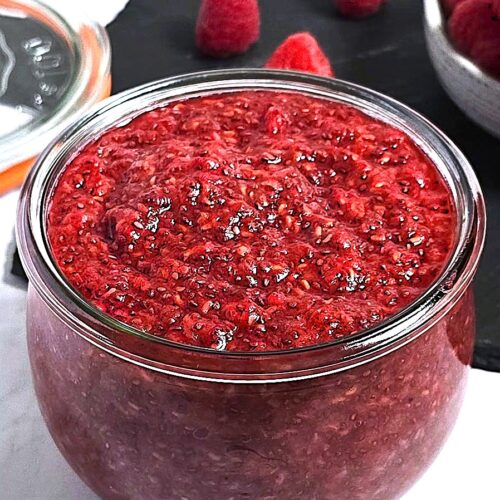 A jar of vibrant pink raspberry chia jam with scattered chia seeds and fresh raspberries.