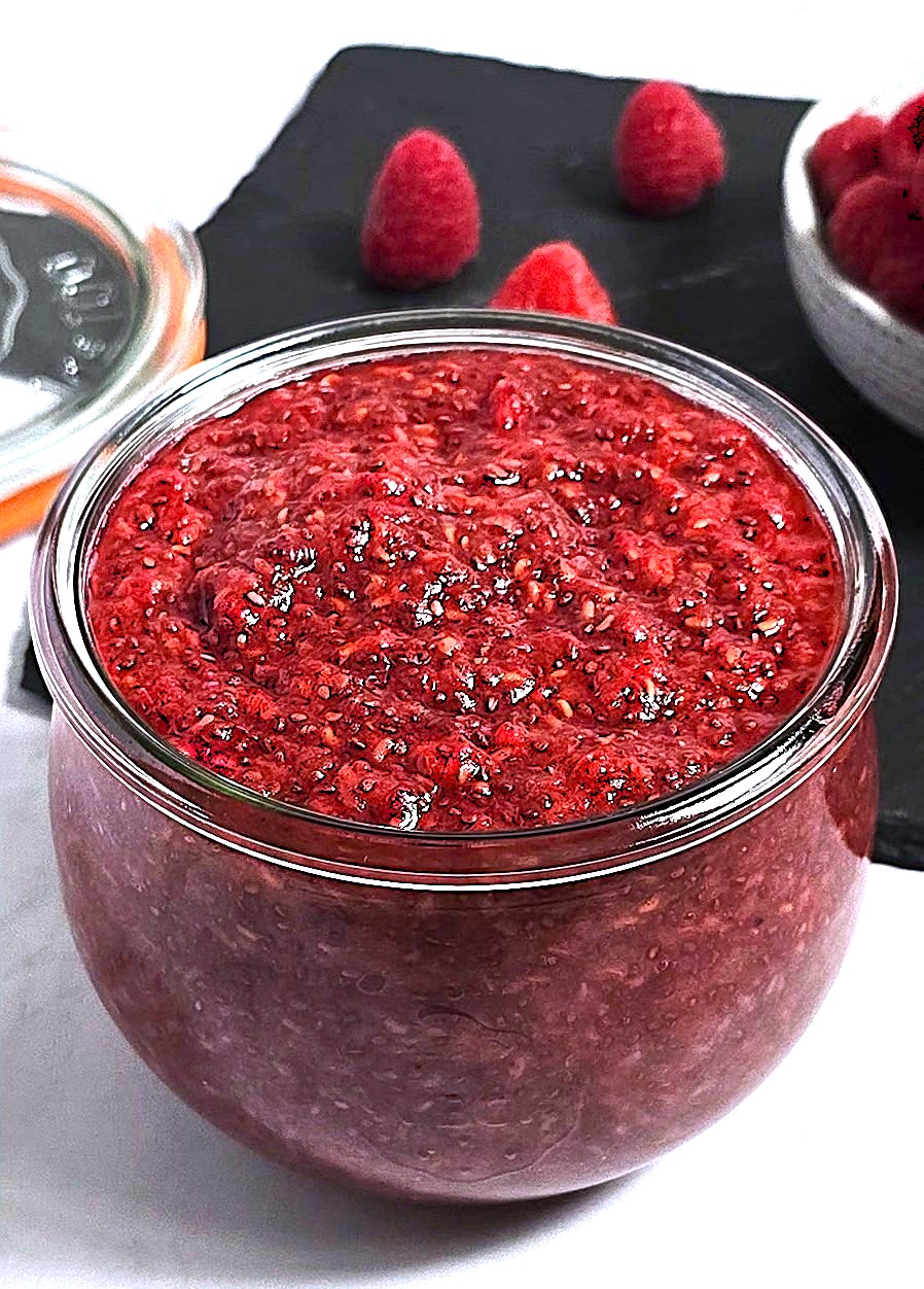 A jar of vibrant pink raspberry chia jam with scattered chia seeds and fresh raspberries.