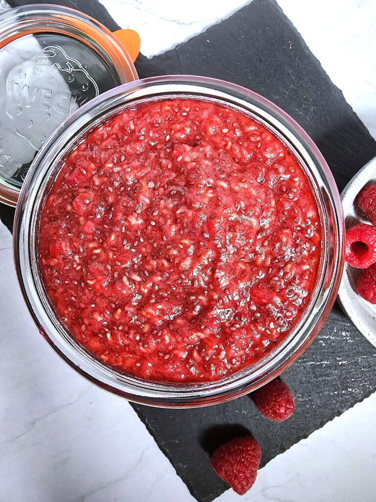 A jar of vibrant pink raspberry chia jam with scattered chia seeds and fresh raspberries.