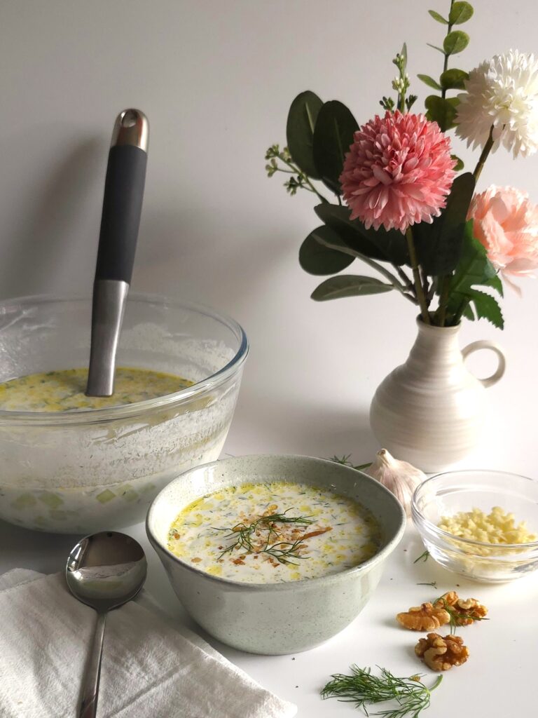 a bowl with Bulgarian tarator along with a bigger bowl, spoon, minced garlic and vase with flowers at the background