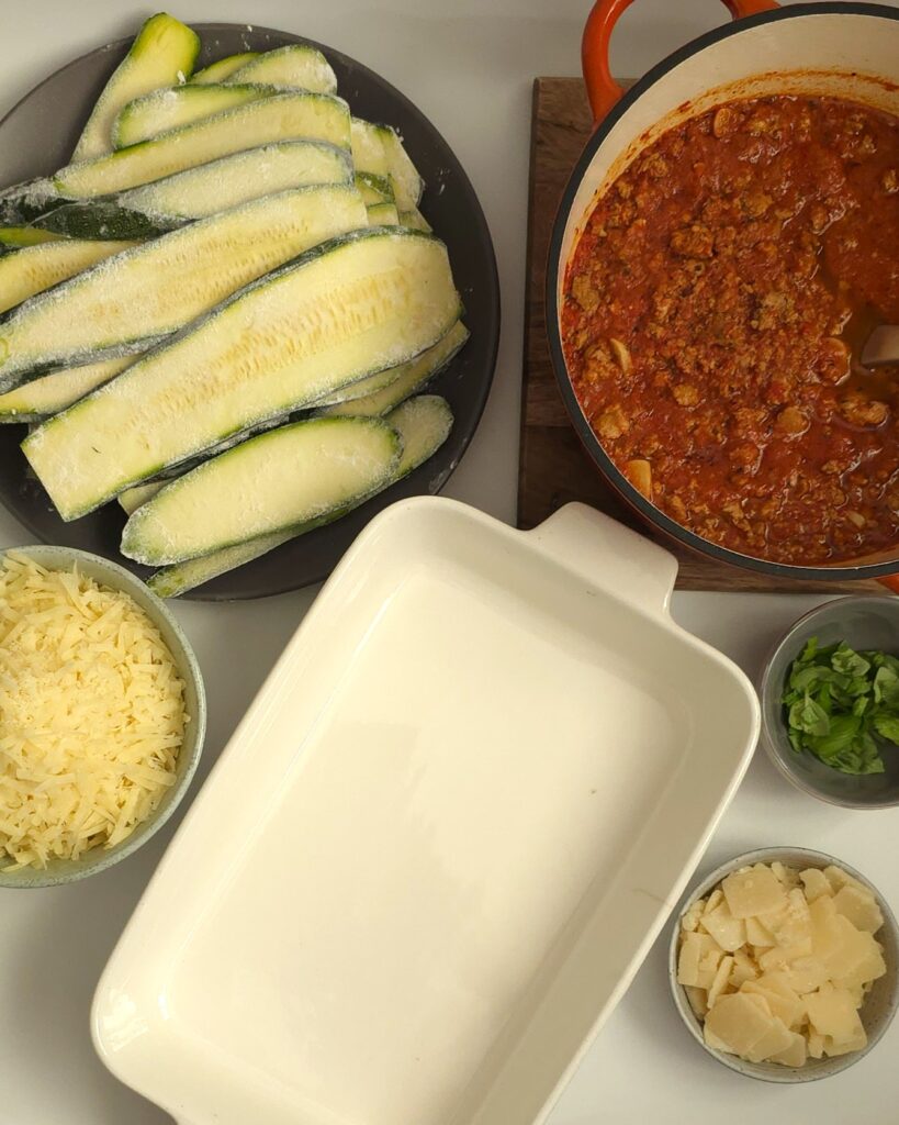 white ceramic baking dish next to floured zucchini slices, grated lactose-free cheese and parmesan