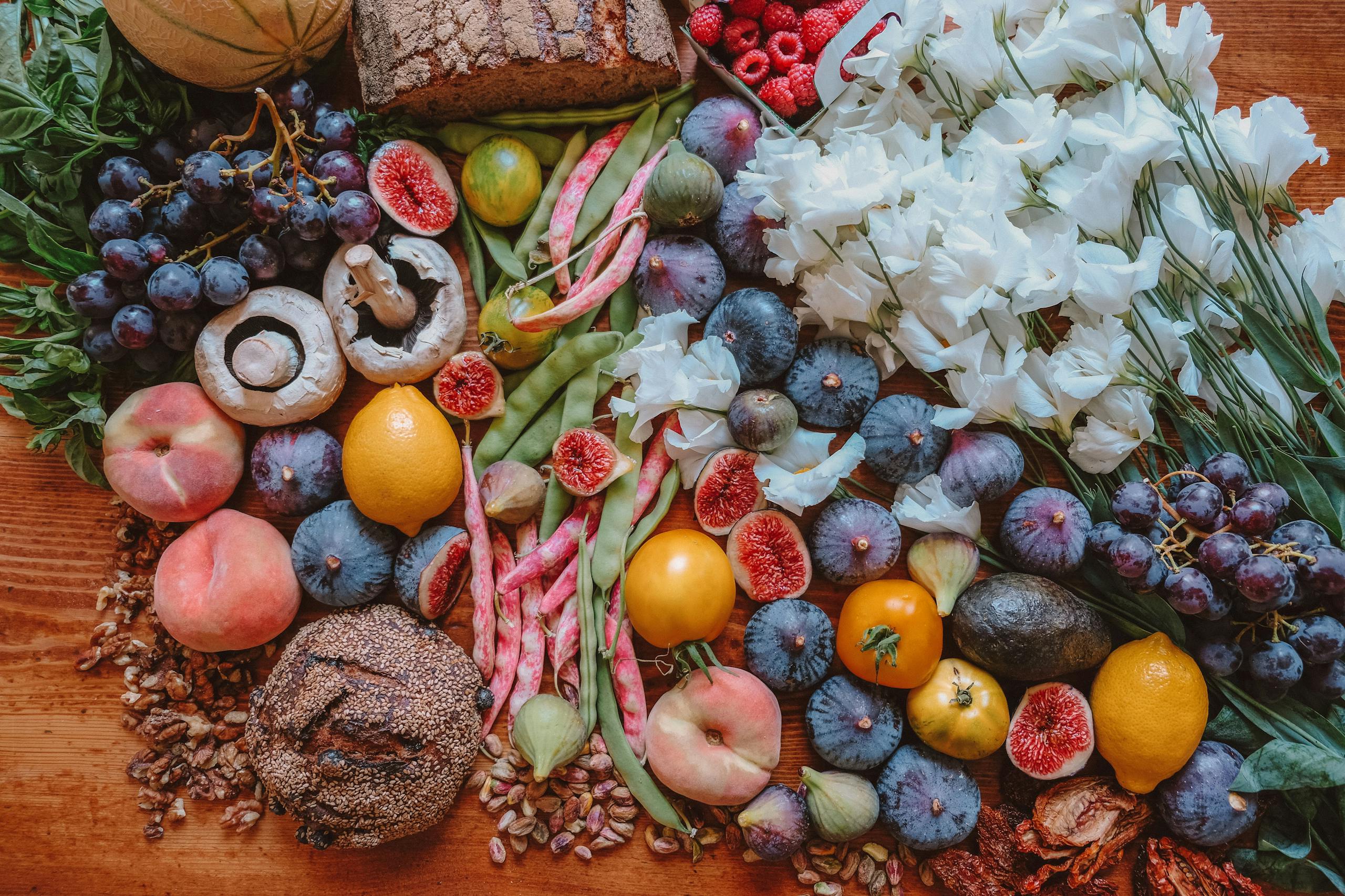 A colorful assortment of fresh fruits, vegetables, bread, and flowers arranged beautifully on a wooden table.