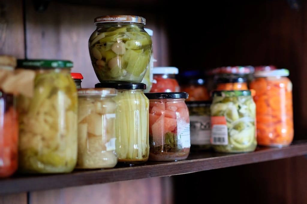 A variety of colorful preserved vegetables in jars arranged on a wooden shelf.