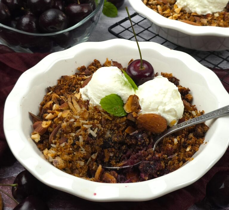 cherry crumble in a white ceramic baking dish next to a bowl of fresh cherries, served with two scoops of strained homemade yoghurt
