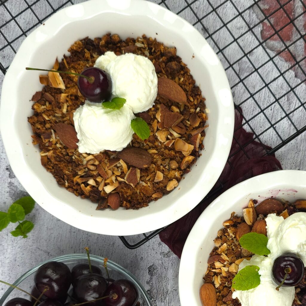 two bowls with cherry crumble in a white ceramic baking dish on a cooling rack, next to a bowl of fresh cherries, served with two scoops of strained homemade yoghurt
