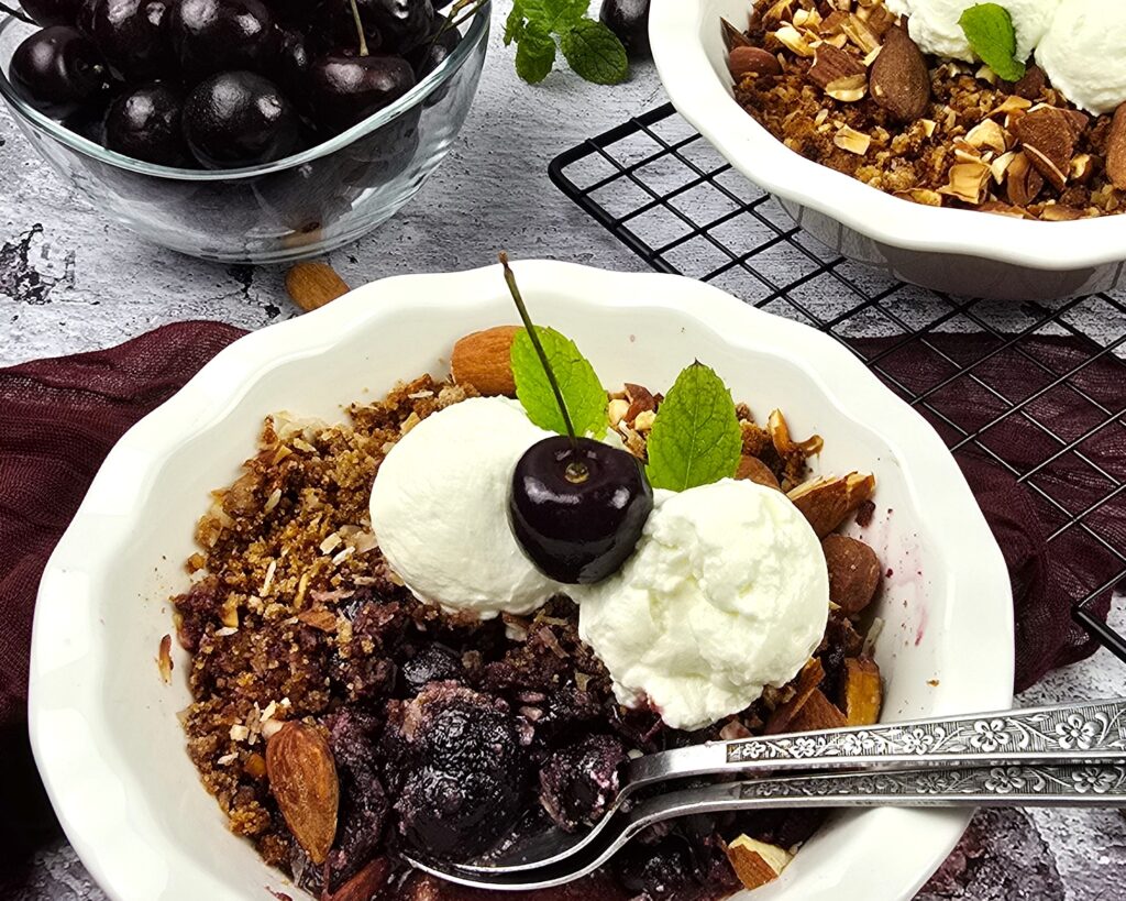 cherry crumble in a white ceramic baking dish next to a bowl of fresh cherries 