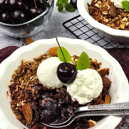 cherry crumble in a white ceramic baking dish next to a bowl of fresh cherries