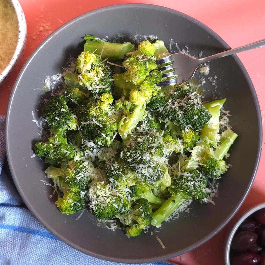 steamed broccoli drizzled with lemon juice and sprinkled with grated parmesan in a dark grey bowl and grated parmesan on top next to bowls with grated parmesan and kalamata olives