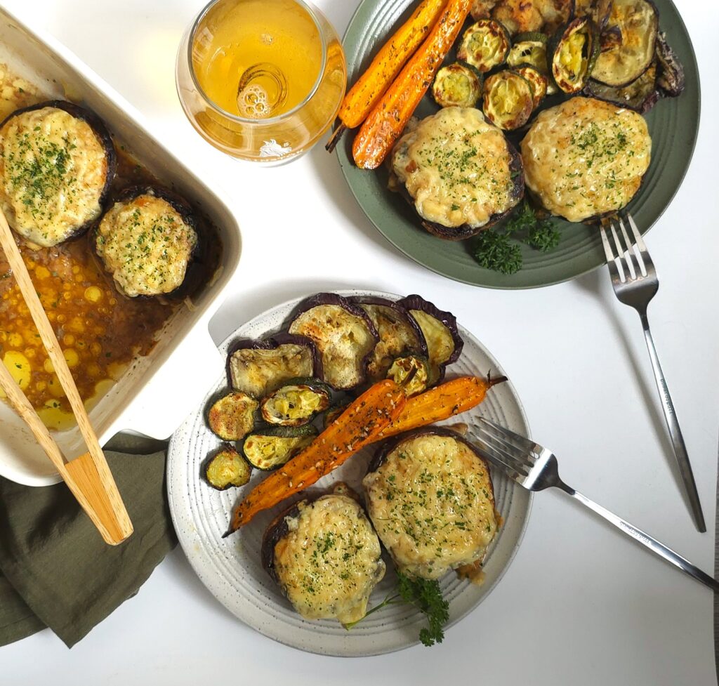 Baked portobello mushrooms stuffed with juicy chicken mince and topped with golden melted cheese, served hot with colourful roasted vegetables in green and grey plates next to white ceramic baking dish.