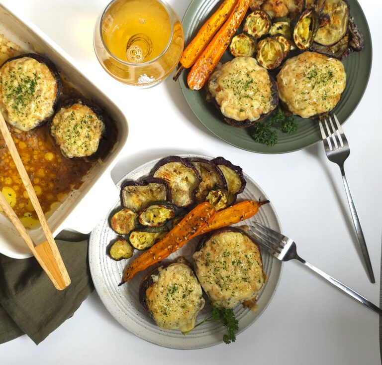 Baked portobello mushrooms stuffed with juicy chicken mince and topped with golden melted cheese, served hot with colourful roasted vegetables in green and grey plates next to white ceramic baking dish.