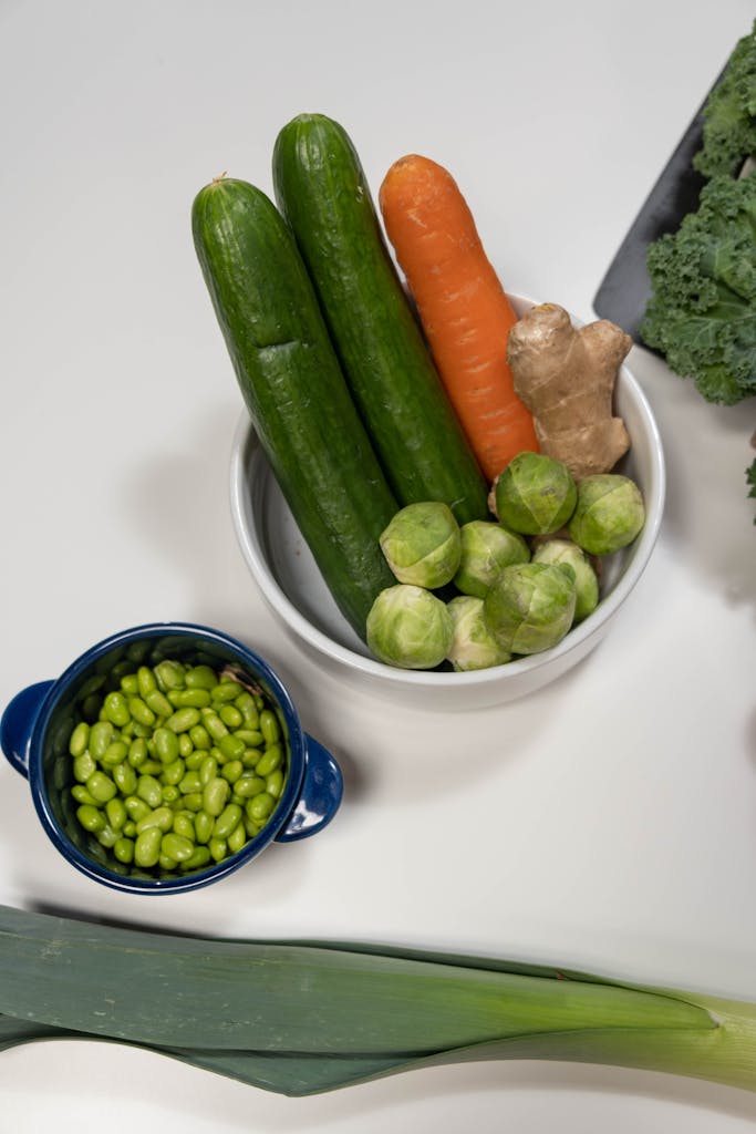 Top view of fibre foods, such as fresh vegetables including cucumbers, carrot, ginger, and edamame on a white surface.