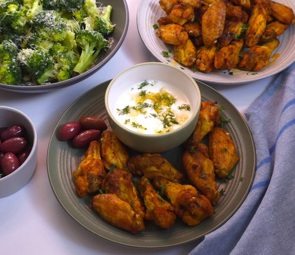 a plate with oven-baked turmeric chicken wings and a bowl with yoghurt dip, next to a plate with broccoli and parmesan