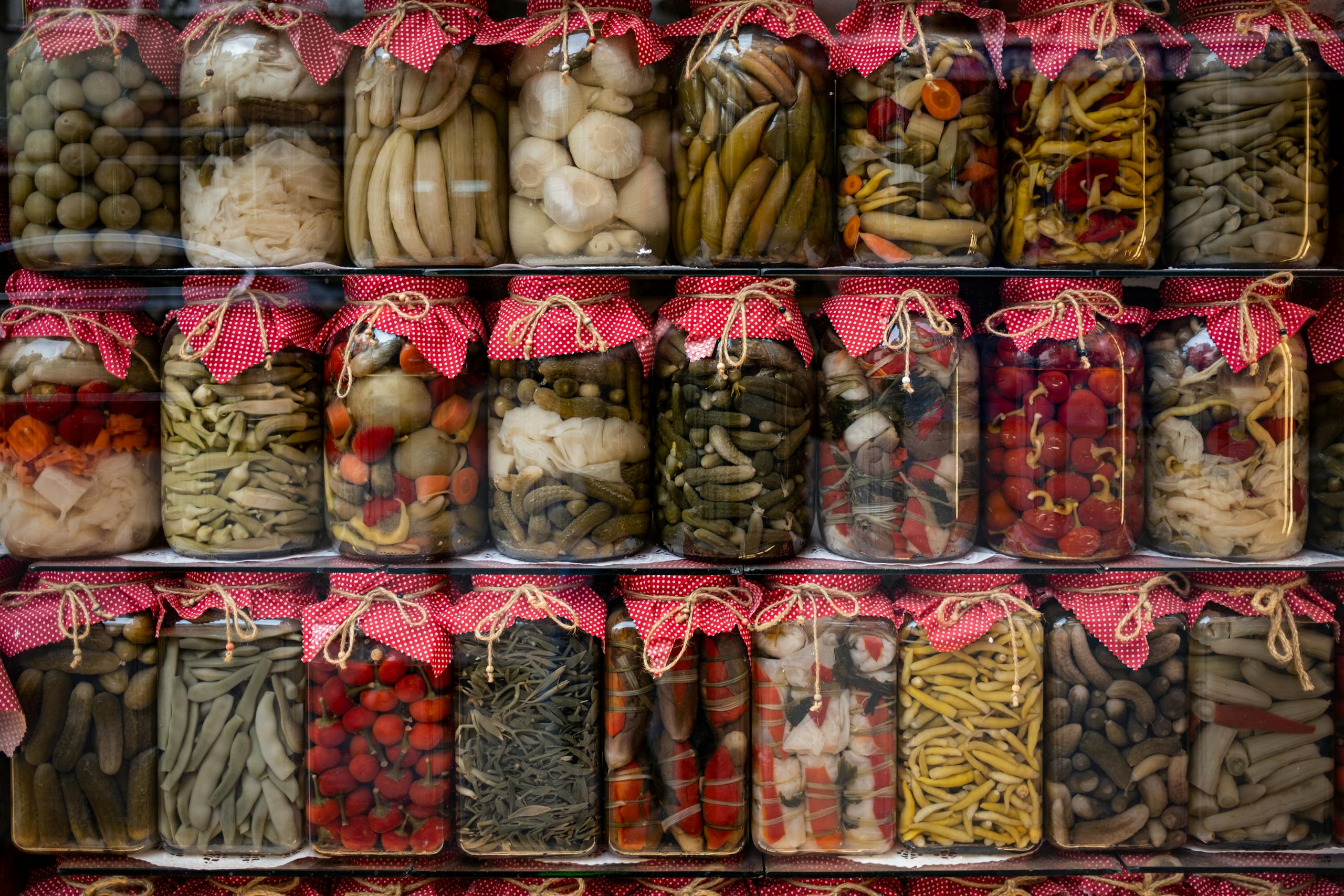 Vibrant assortment of pickled vegetables in jars with red fabric covers, displayed on shelves.