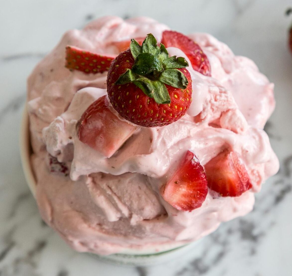 A bowl of homemade strawberry ice cream with fresh strawberries on a marble countertop.