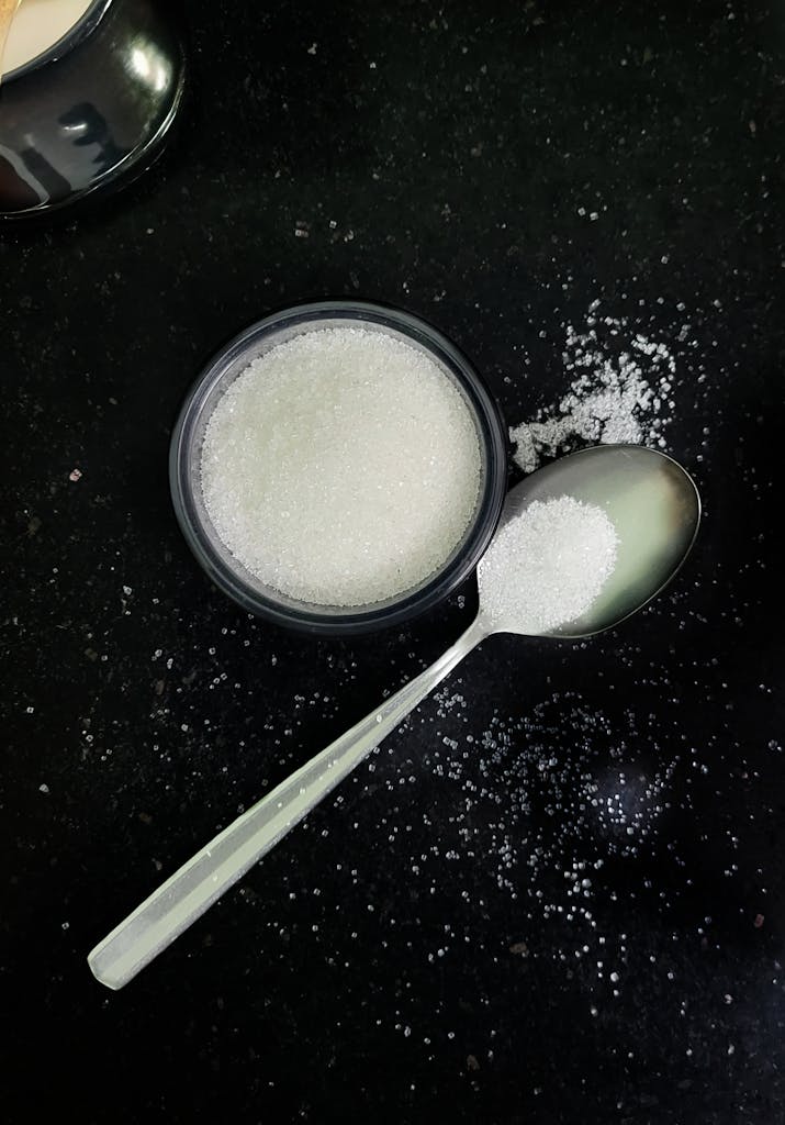 A spoon with granulated sugar beside a full bowl on a black countertop. Simple yet elegant kitchen scene.