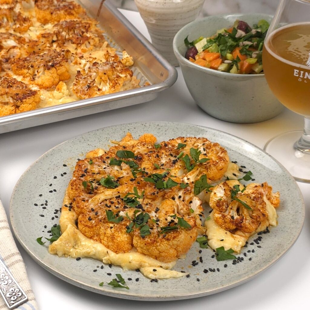 a cauliflower steak in a green stoneware plate next to a bowl with a salad and roasting tray 