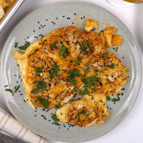 a golden baked cauliflower steak in a plate sprinkled with fresh parsley and black sesame