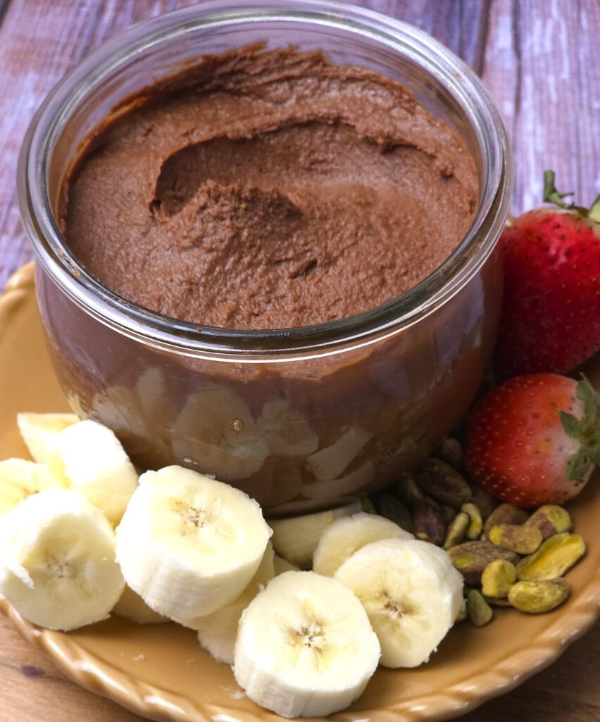 a glass jar with cocoa hummus recipe next to sliced banana, pistachio and strawberries