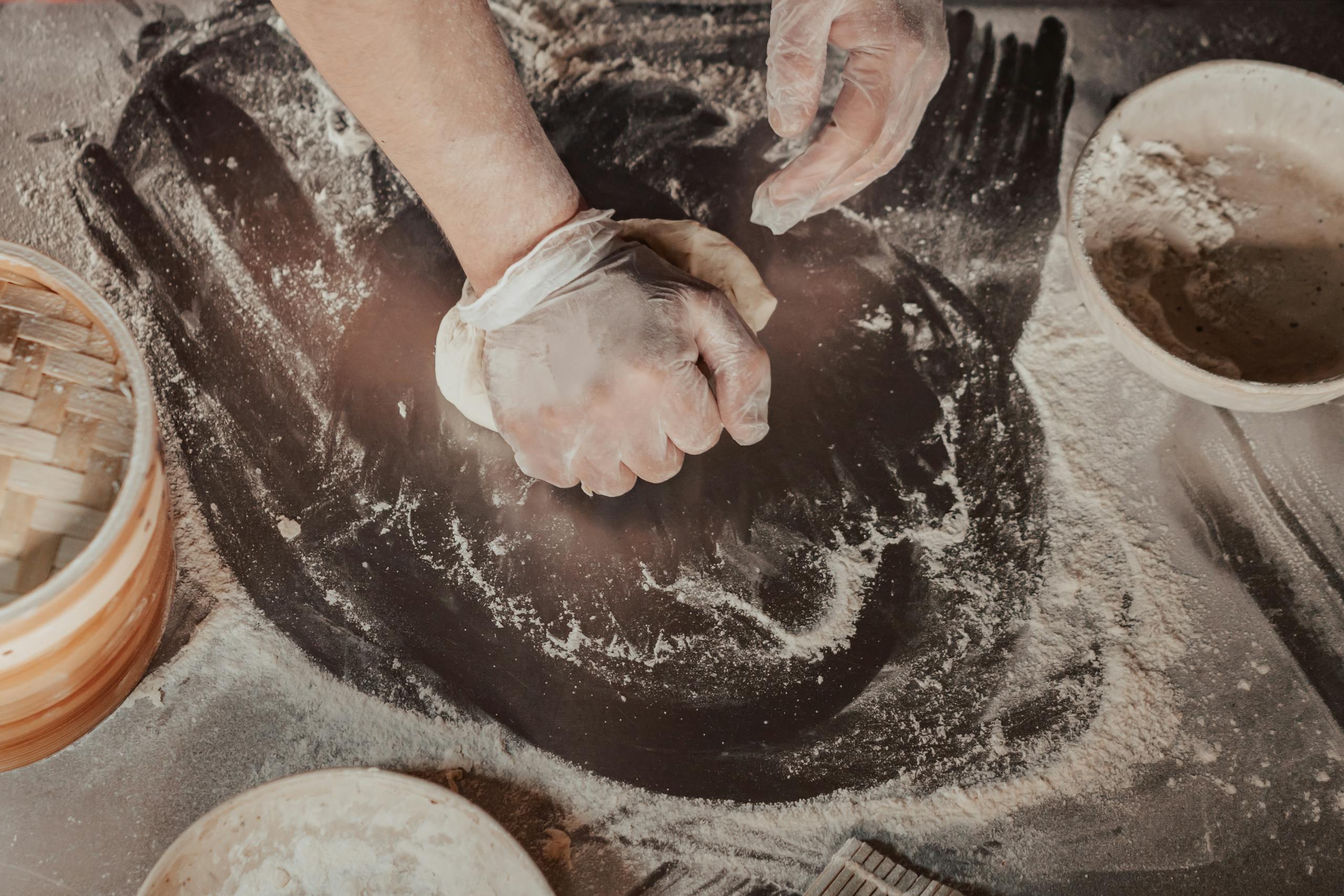gluten-free bake scene - hands in gloves kneading dough on a floured table with bowls and baking tools nearby.
