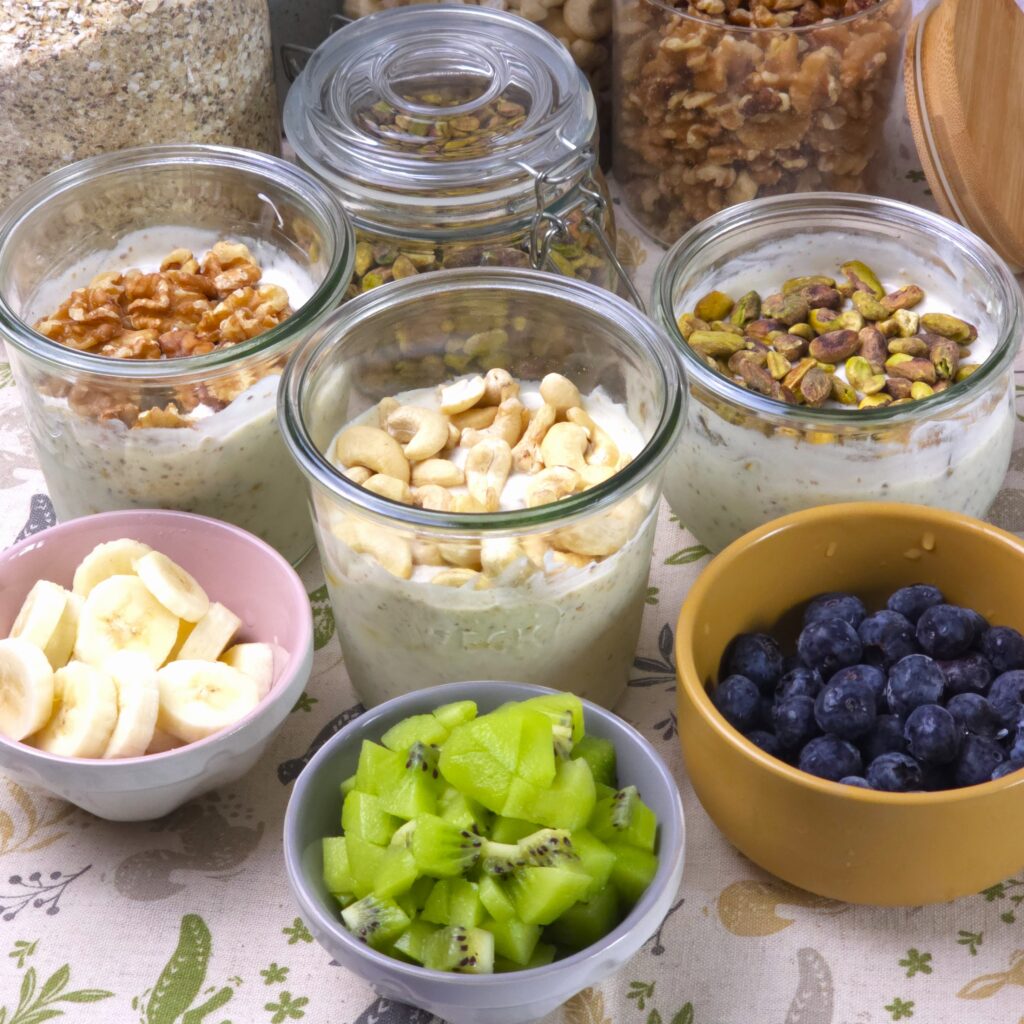 three jars with gluten-free overnight oats, topped with walnuts, cashew and pistachio next to three bowls with kiwi, banana and blueberries