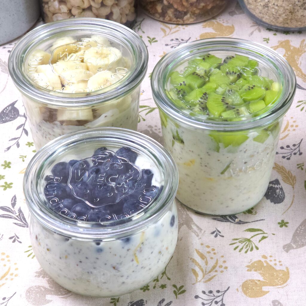 Gluten-free overnight oats layered with yoghurt, fruit, and nuts in glass jars with lids, topped with blueberries, kiwi, and banana.