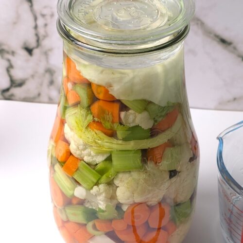 A jar with chopped carrots, cauliflower and celery sticks in a brine