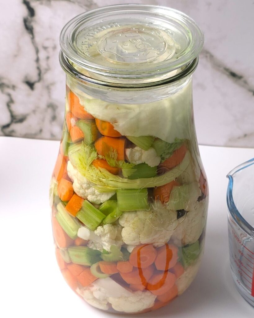 A jar with chopped carrots, cauliflower and celery sticks in a brine