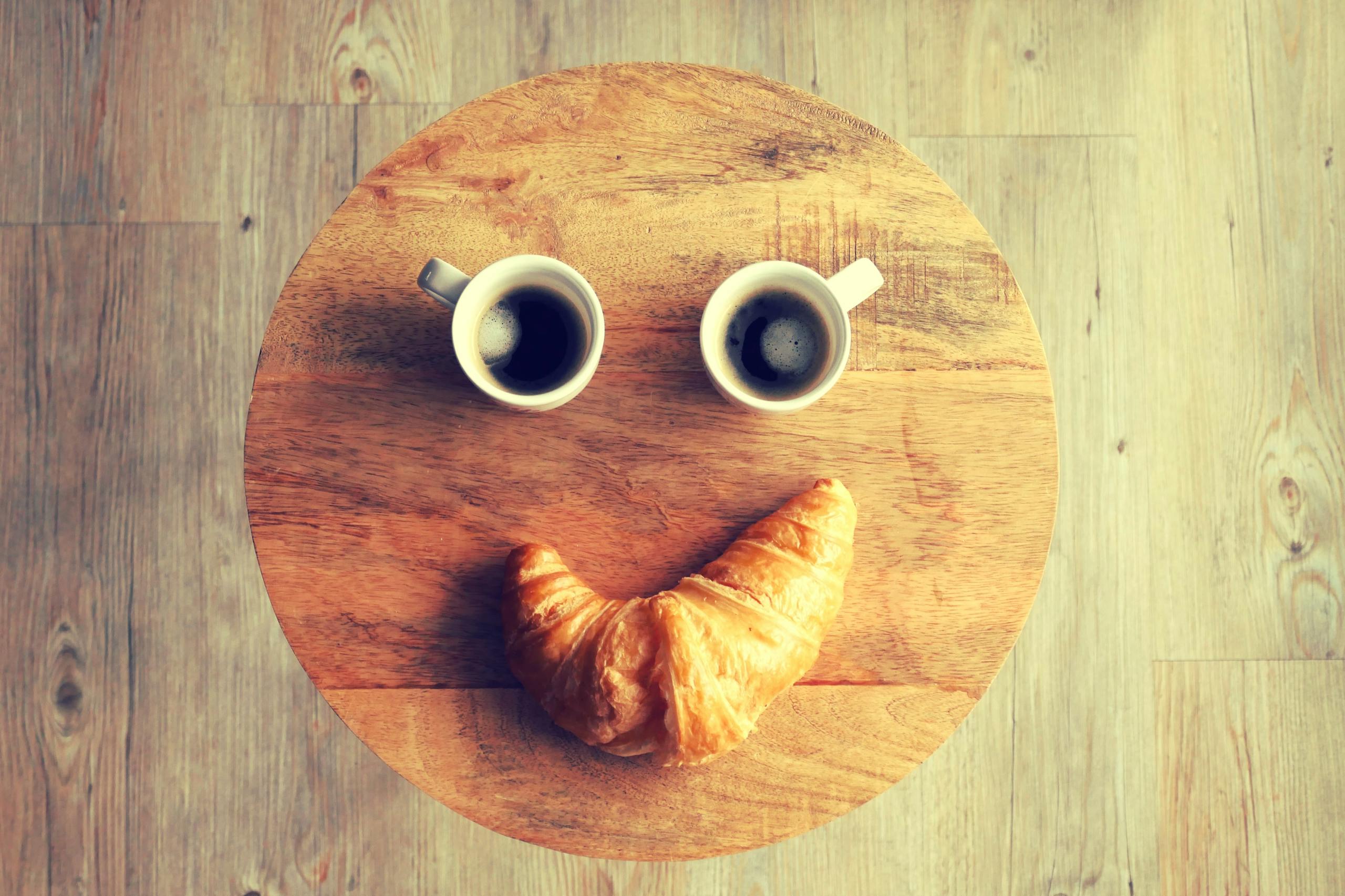 Top view of coffee cups and croissant arranged humorously as a smiley face on a wooden table.
