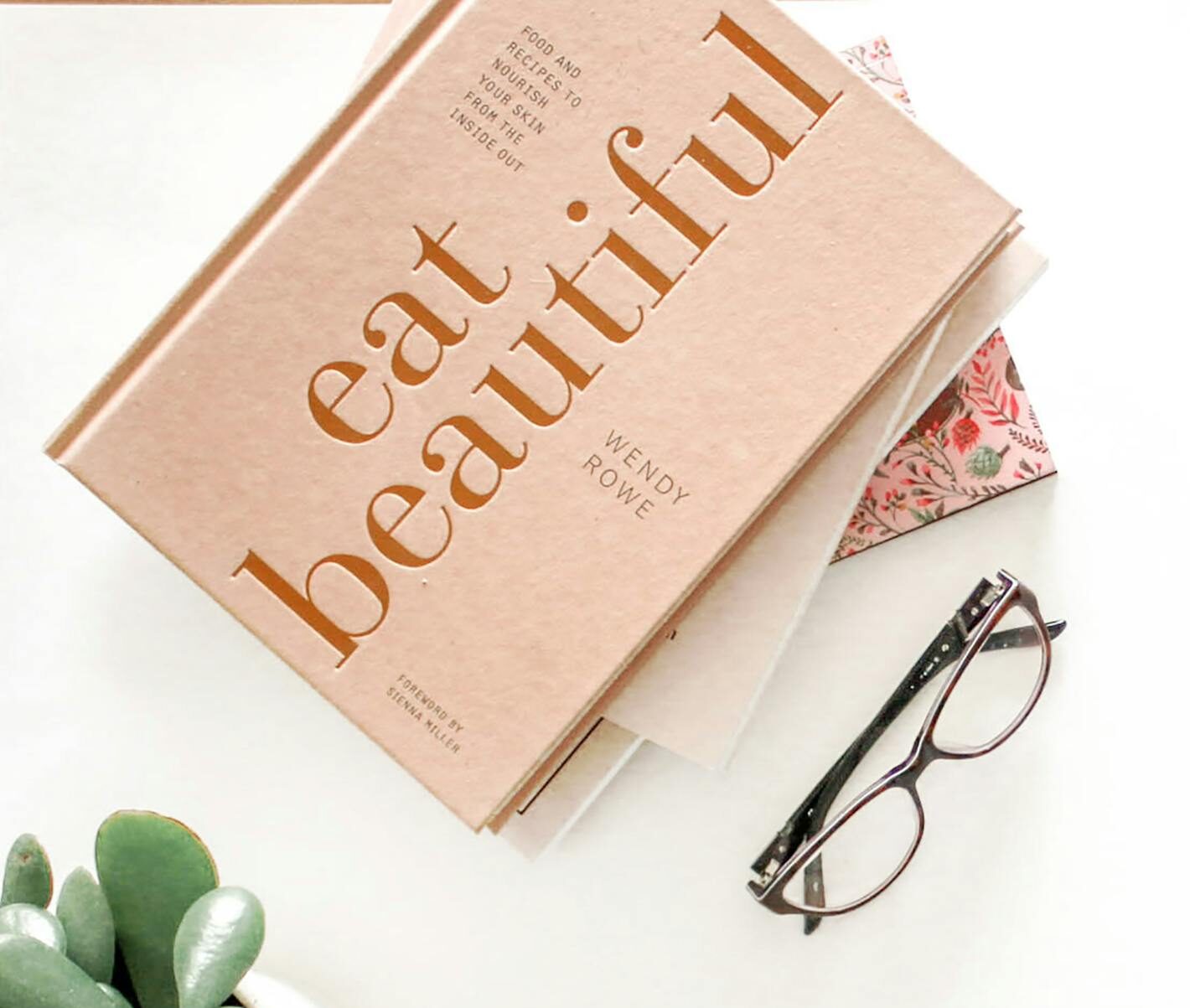 A minimalist flatlay featuring a book, eyeglasses, and a plant on a white background, while writing book reviews.
