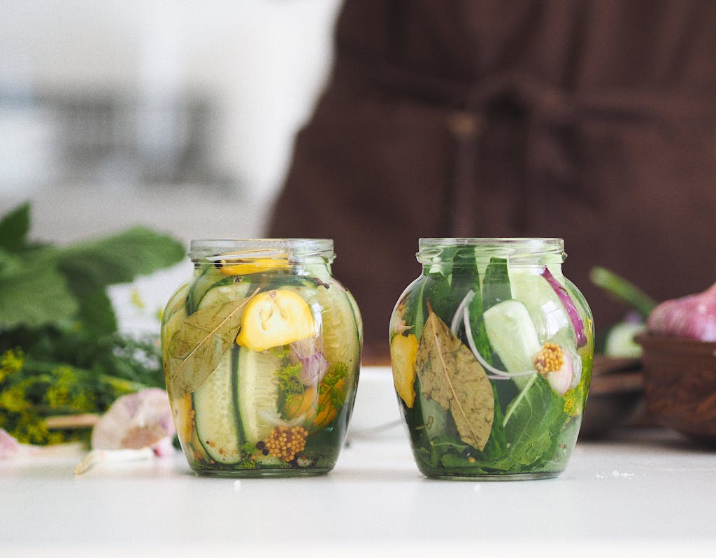 Two glass fermentation jars filled with homemade pickled cucumbers and herbs on a white kitchen surface.
