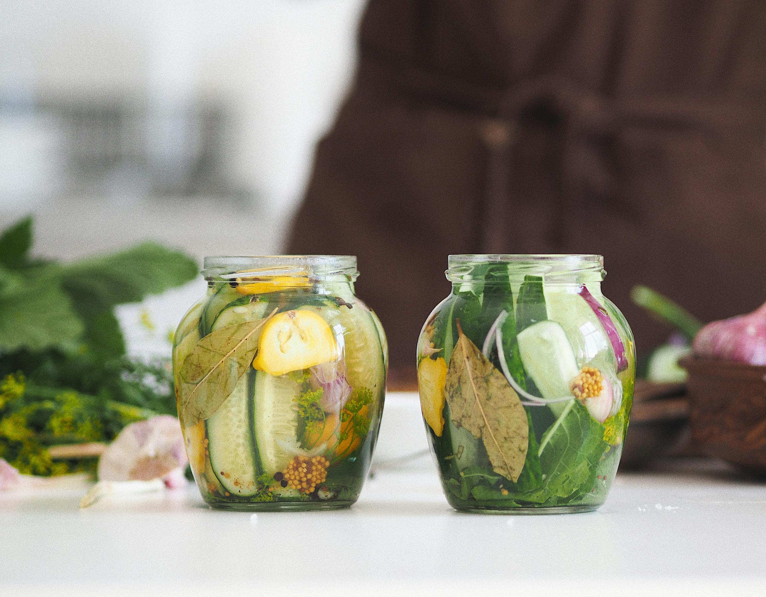Two glass fermentation benefits jars filled with homemade pickled cucumbers and herbs on a white kitchen surface.