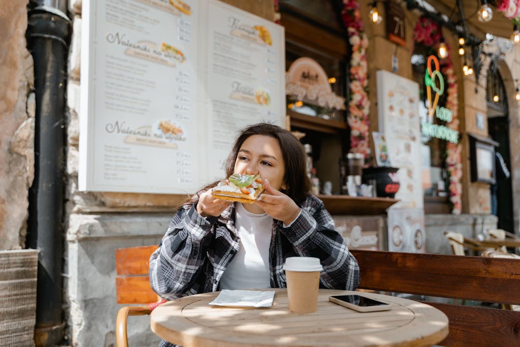 Woman in plaid shirt enjoying a sandwich at an outdoor cafe with a coffee.