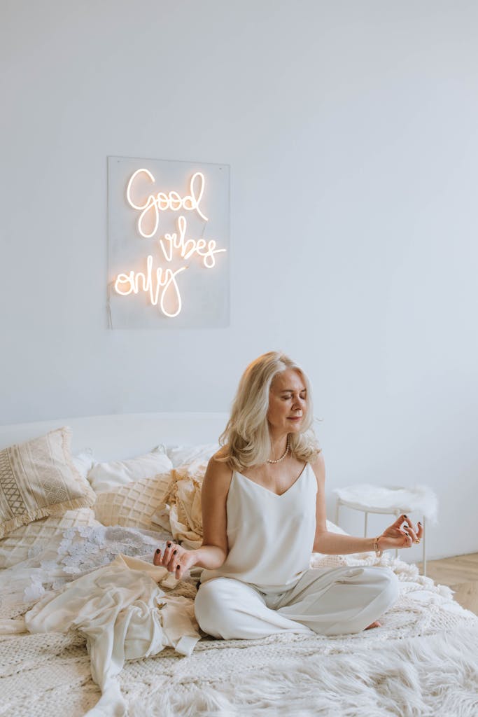 Serene woman meditating on a cozy bed under 'Good Vibes Only' neon sign.