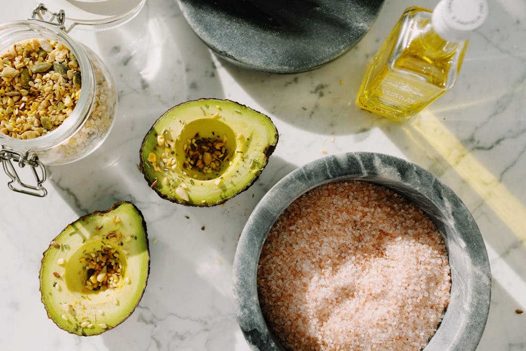 Overhead view of avocado halves, seeds, pink salt, and olive oil on marble surface.