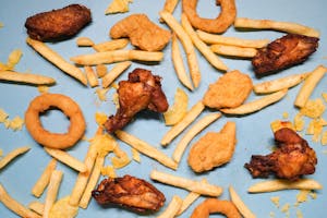 Ultra-processed foods - from above of french fries and grilled chicken wings with golden deep fried onion rings and nuggets with chips on blue background in bright studio