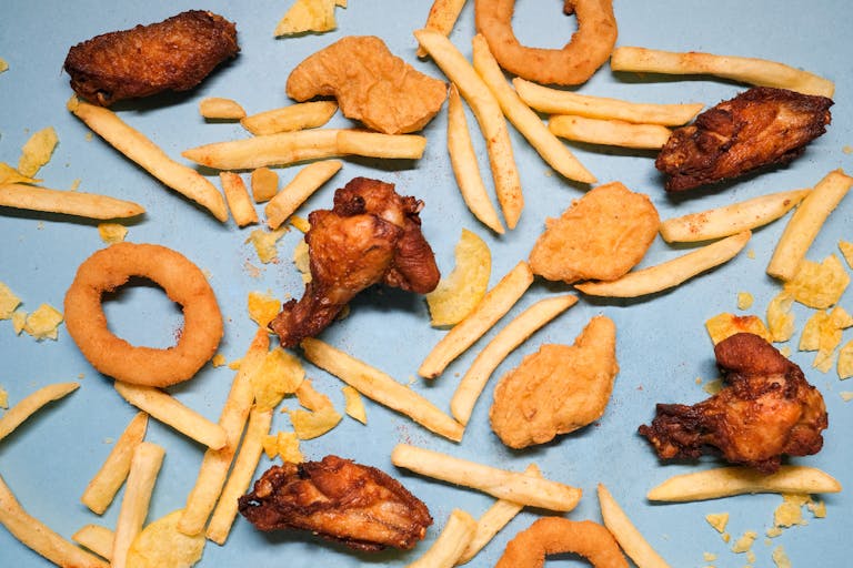 Ultra-processed foods - from above of french fries and grilled chicken wings with golden deep fried onion rings and nuggets with chips on blue background in bright studio