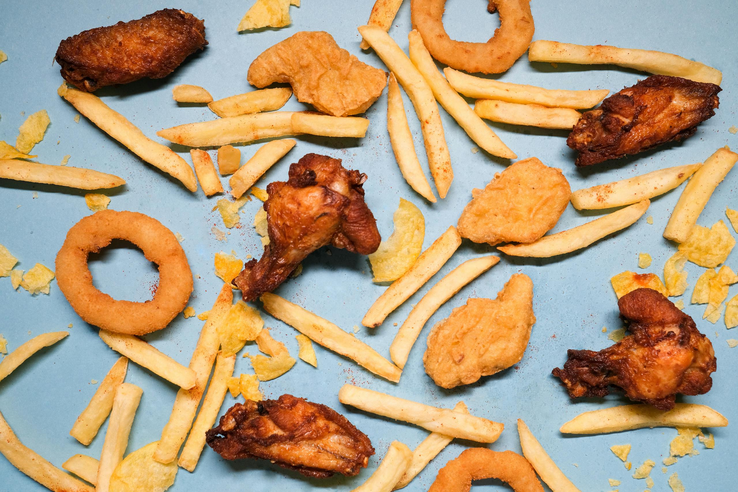 Ultra-processed foods - from above of french fries and grilled chicken wings with golden deep fried onion rings and nuggets with chips on blue background in bright studio