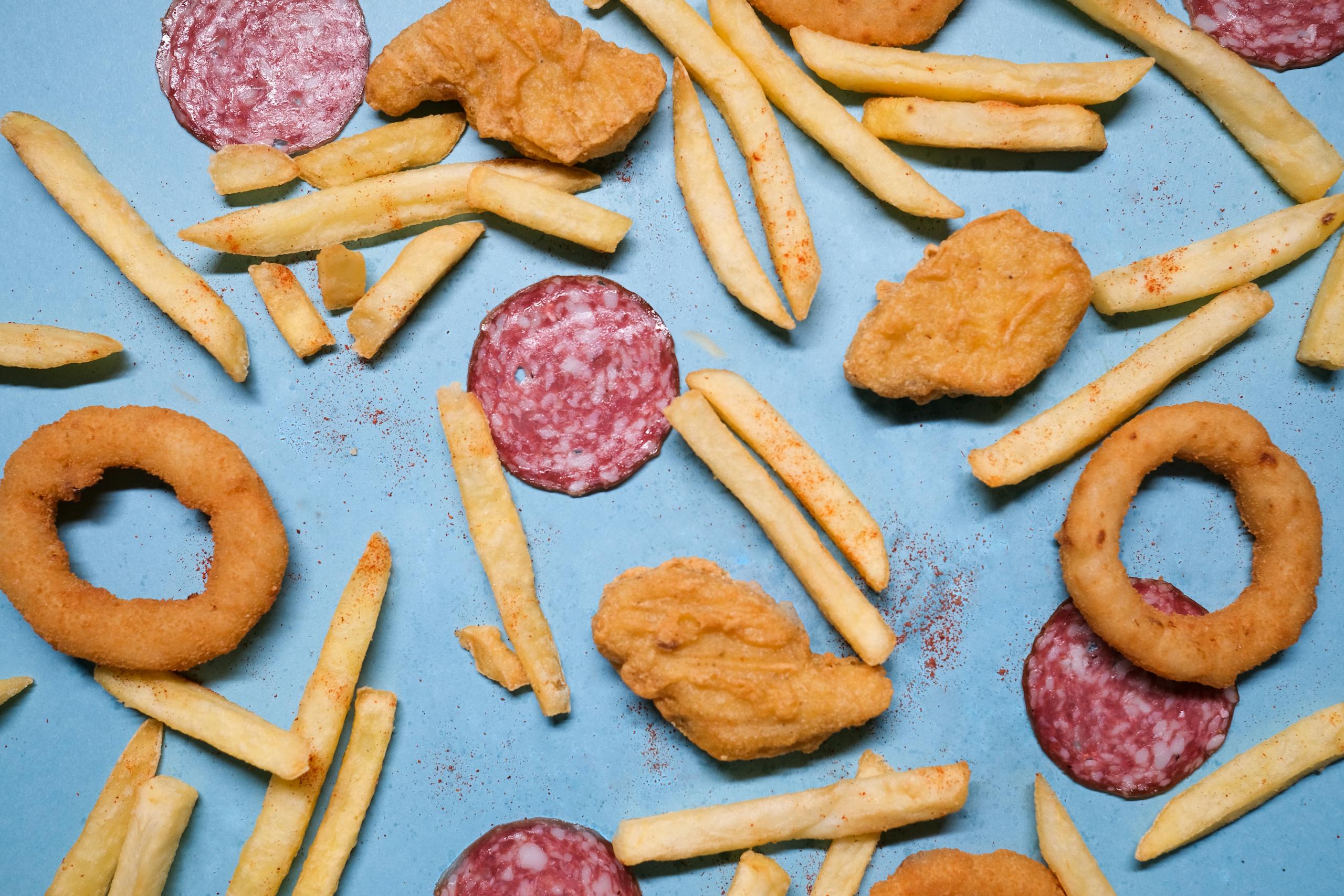 From above of salami slices with fried potatoes and deep fried onion rings with golden chicken nuggets on blue background in light studio