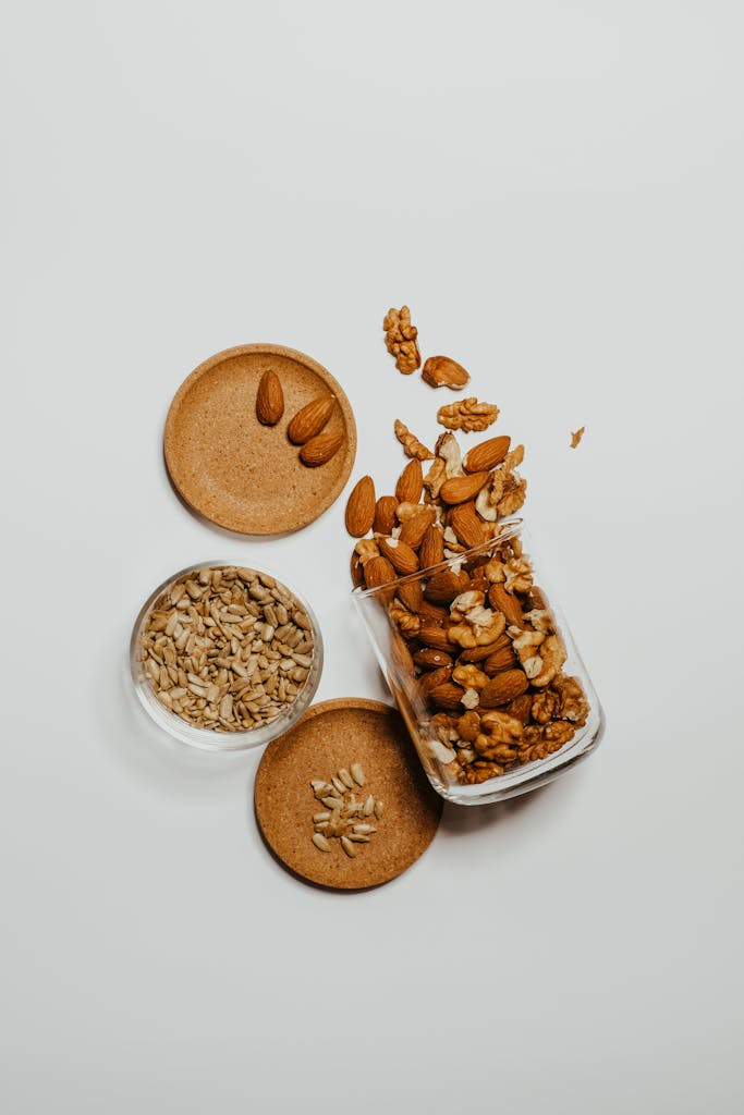 A flat-lay of mixed almonds, walnuts, and sunflower seeds in glass jars over a white surface.