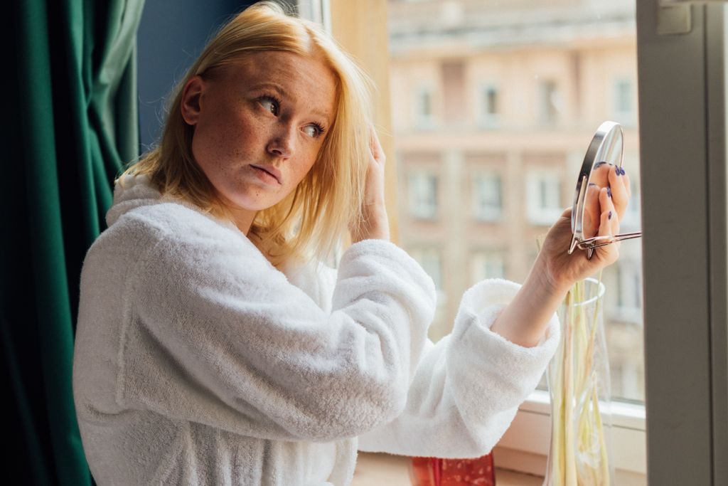 A woman in a white bathrobe looking in a mirror by the window.