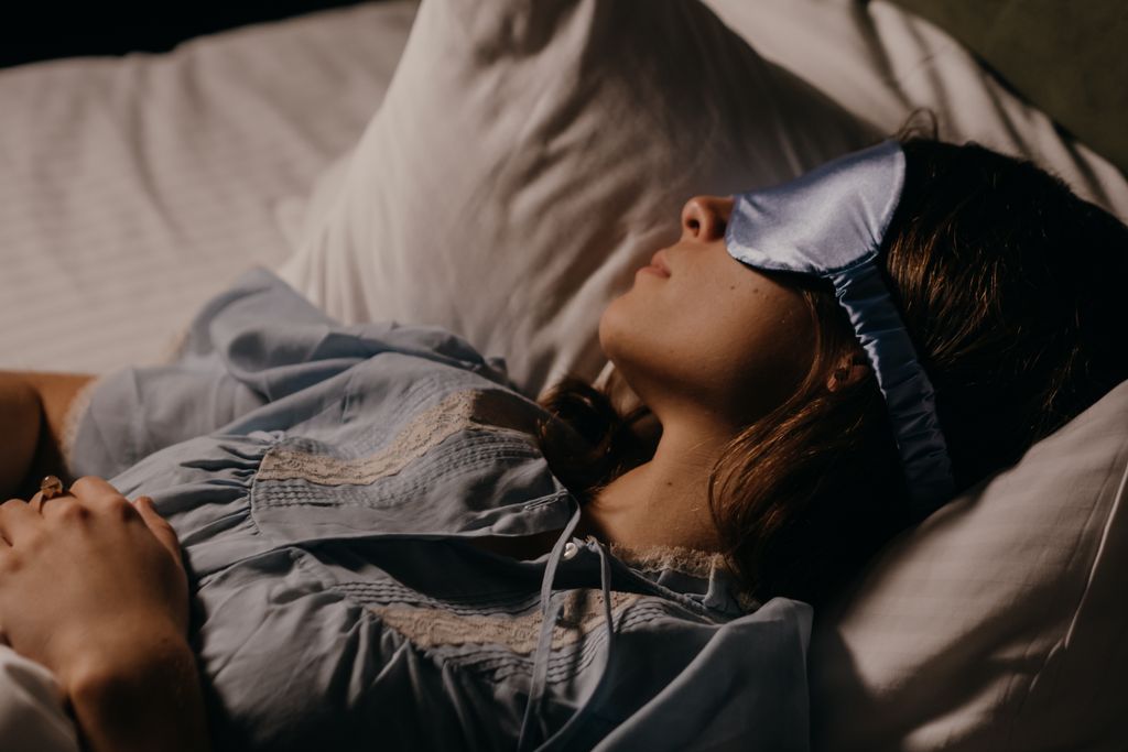 A woman peacefully sleeping with a blue eye mask in a dimly lit room.