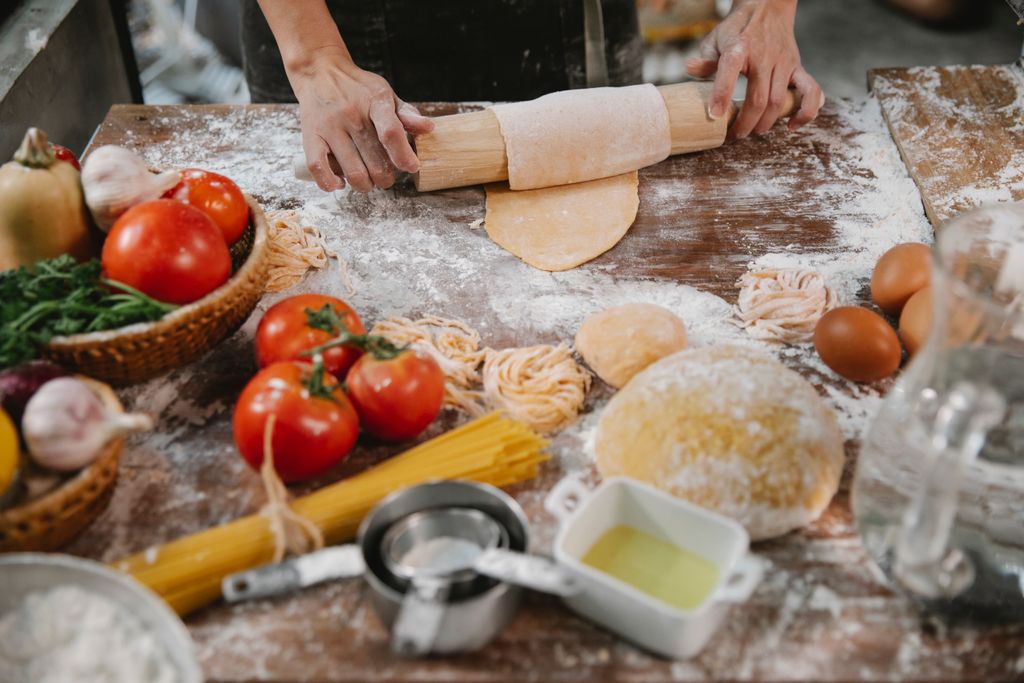 Crop anonymous female cooking homemade pasta on table covered with flour among vegetables