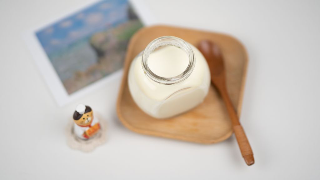 From above arrangement of fresh milk food matrix in Bulgarian yogurt in glass jar placed on wooden saucer near funny cat figurine and blurred photo of nature