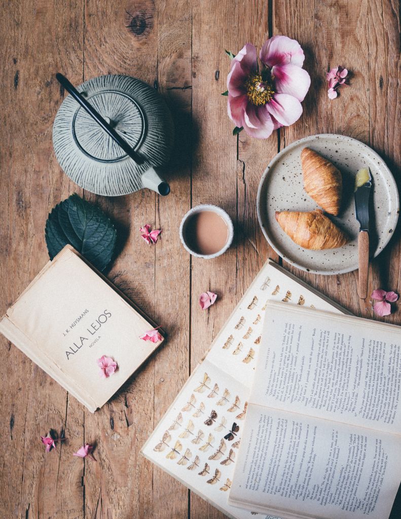 Top view of opened books placed on wooden table near kettle with cup of coffee served near plate with croissants and bright flower
