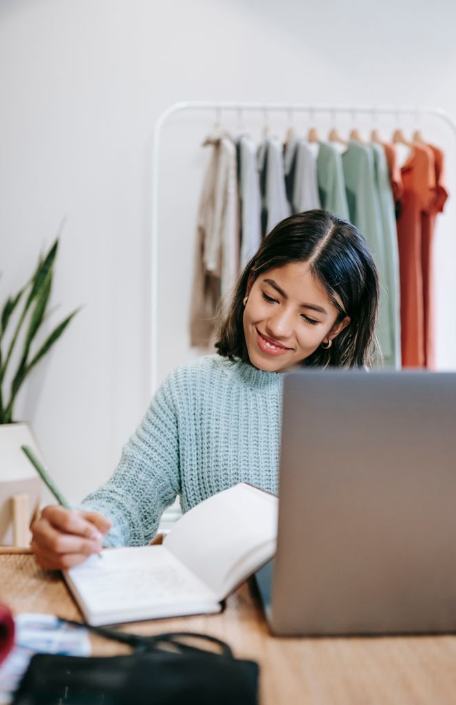 Cheerful Latin American female remote worker with agenda and netbook working at table in house